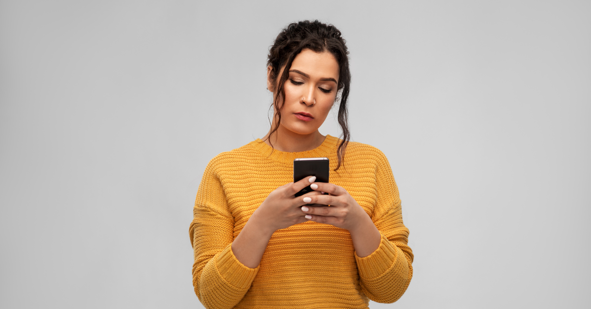 Na obrázku je žena s mobilom v ruke, ktorá sa naň pozerá so zmesou smútku a zamyslenia.